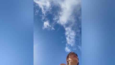 Video of a young man's face looking upward at a bright blue sky filled with fluffy white clouds. His hair is short and light brown, and he is wearing a light-colored shirt.