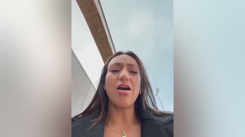 A video of a woman with medium brown skin, long dark hair, and a black jacket, captured from a low angle, showing her from the chest up. She appears to be in a moment of intense emotion.
