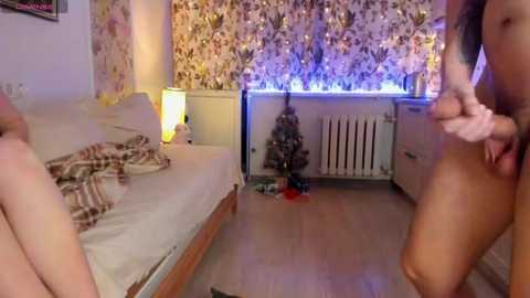 Media: A video of a cozy, dimly lit bedroom with a naked woman in the foreground, a bed with a plaid blanket, a floral-patterned wall, and a Christmas tree.