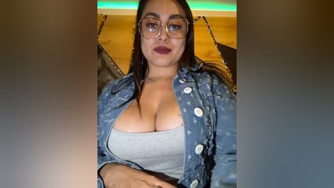 Video of a plus-size Latina woman with medium skin tone, long dark hair, and large breasts, wearing a denim jacket over a low-cut grey top, sitting in a dimly lit room with green LED lighting in the background.