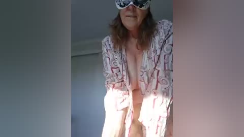 Video of a middle-aged woman with shoulder-length brown hair, wearing a low-cut, patterned robe, a white blindfold, and a playful expression. Background shows a simple, dimly lit room with light-colored walls.