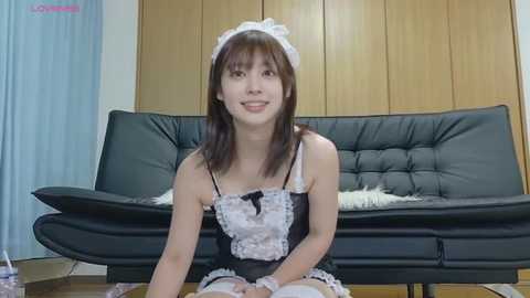 Media: Video of an East Asian woman in a revealing maid outfit with white lace and black satin, sitting on a black leather couch. Background includes light blue curtains and wooden cabinets.