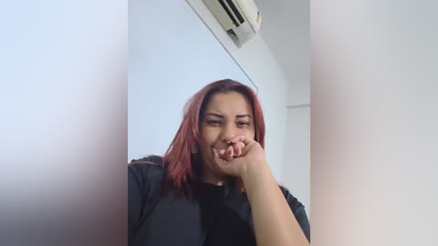 Video of a young woman with shoulder-length, dyed red hair, wearing a black shirt, covering her mouth in a gesture of shock or surprise, against a white wall with an air conditioning unit visible.