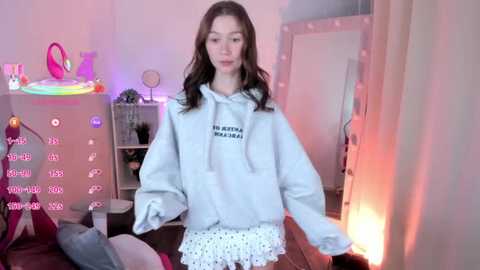 A video of a young, slender, Caucasian girl with long, wavy brown hair, wearing a gray hoodie and white shorts, standing in a dimly lit, pink-lit bedroom with various toys and a soft, plush animal.