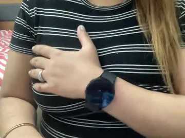 Video of a woman with light skin, wearing a black and white striped shirt, placing her left hand on her chest. She has long blonde hair and a black smartwatch on her left wrist. Background includes a red and white patterned bedspread.