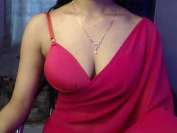 Video of a woman wearing a revealing, vibrant pink dress with thin straps, revealing ample cleavage. She has a gold necklace with a heart pendant. Background is dimly lit with indistinct furniture.