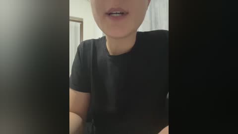 Video of a light-skinned person with straight, dark hair, wearing a black t-shirt, partially obscured by a dark vertical shadow on the left. Background includes a framed picture and white wall.