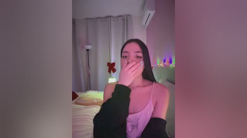 Media: Video of a young woman with long black hair, wearing a light pink tank top and black jacket, covering her face with her hand, in a dimly lit bedroom with a bed, red floral decor, and string lights.