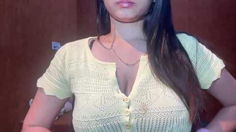 Video of a woman with long, straight brown hair, wearing a cream-colored, crocheted, short-sleeved blouse with a deep V-neck, revealing cleavage. She stands in front of a wooden door, with a simple necklace around her neck.
