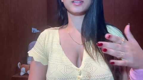 Video of a light-skinned woman with long, straight black hair, wearing a yellow crochet top, and red-painted nails, smiling slightly. Background shows wooden paneling and a stuffed animal.