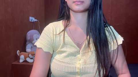 Video of a young woman with long, straight black hair, wearing a yellow knit top and a necklace, standing in a room with wooden walls and a stuffed animal.