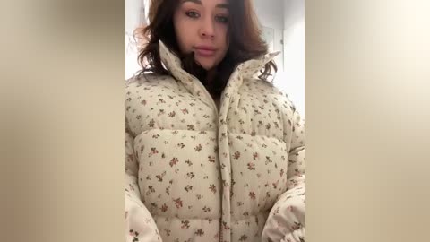 A video of a young woman with long, wavy brown hair, wearing a puffy, beige jacket with floral patterns. She has a neutral expression and is indoors, with a blurred background.