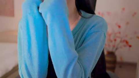 A video of a person wearing a turquoise sweater, holding a blue towel over their head in a softly lit, pastel-colored bedroom with floral wallpaper.