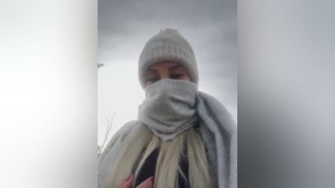 Video of a person with light skin wearing a gray beanie, gray mask, and a light-colored scarf, standing indoors in a narrow corridor with a blurry, overcast background.