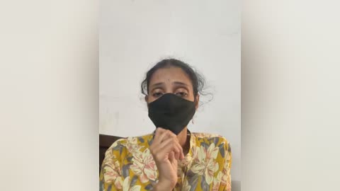Video of a South Asian woman with medium brown skin and dark hair tied back, wearing a floral-patterned blouse, black face mask, and looking concerned, standing in a white-walled room.