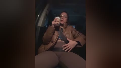 Media: Video of a woman in a car, wearing a brown jacket and glasses, with a belt across her waist, holding a white cup, dim lighting, and blurred background.