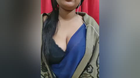 Video of a South Asian woman with medium skin tone, wearing a blue saree with a black lace bra, gold earrings, and a beige jacket with floral pattern. Background shows a red curtain.