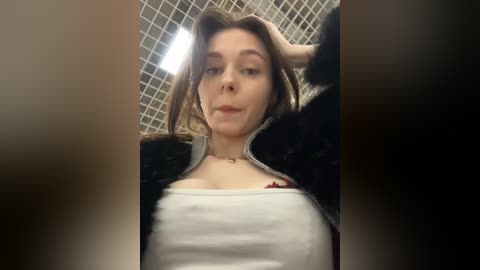 A candid video of a young Caucasian woman with light skin and shoulder-length brown hair, wearing a white tank top and black jacket, standing indoors near a tiled wall.