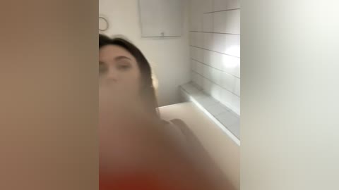 A blurry video of a woman with shoulder-length brown hair and a light complexion, partially obscured by a shower curtain, standing in a modern bathroom with white tiles and a window.