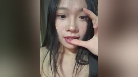 Media: Video of an Asian woman with long black hair, fair skin, and light makeup, biting her lower lip. She wears a black top. The blurred background suggests a private indoor setting.