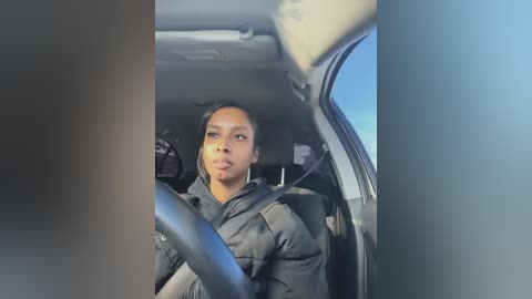 Media: A video of a Black woman with medium brown skin, wearing a black puffy jacket, sitting in a car with the seatbelt on, looking serious.