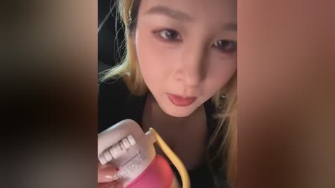 Video of an Asian woman with long blonde hair, fair skin, and brown eyes, holding a pink and yellow medicine cup, wearing a black top, in a blurred, dimly lit room.