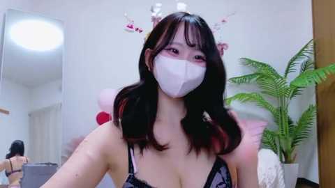 Video of an East Asian woman with long, straight black hair, wearing a white face mask and a black lace bra. She stands in a room with a mirror, potted plant, and pink decor.
