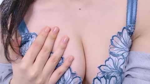 Video of a light-skinned woman with medium-sized breasts, wearing a lacy blue bra with floral embroidery. Her hand, with neatly trimmed nails, gently cups her left breast. Background is blurred, focusing attention on her chest and hand.