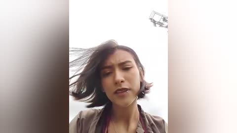 Video of a young woman with shoulder-length dark hair, wearing a brown top, facing the camera with a neutral expression, set against a bright, diffused background.