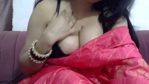 Video of a South Asian woman with medium skin tone and long black hair, wearing a pink sari, partially covering her black bra, revealing her large breasts. She's on a pink couch, wearing a gold bracelet.