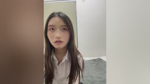 A video of a young Asian woman with long, straight brown hair, wearing a white shirt, standing in a beige room with a grey carpet.