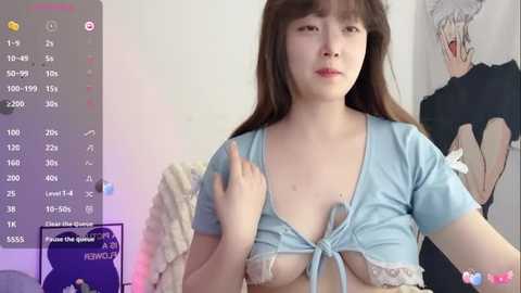 A video of an Asian woman with long brown hair, wearing a light blue crop top, partially exposing her breasts, in a bedroom with anime posters and a bed in the background.