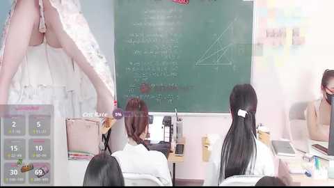 Media: Video of a classroom scene: a teacher in a white dress and a student in a ponytail sit at a desk, while another student in a tank top looks at a green chalkboard.