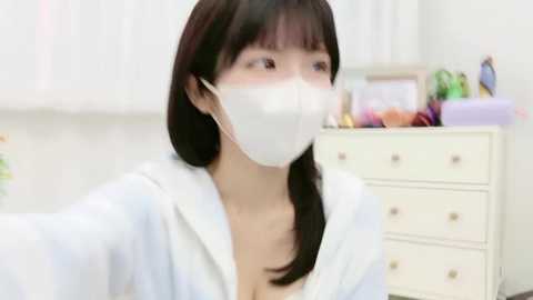Video of an Asian woman with straight black hair, wearing a white mask, light blue jacket, and sitting in a brightly lit room with white furniture and a desk filled with colorful items.