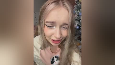 Video of a young woman with long, wavy blonde hair, wearing a white shirt with a black cow print, looking towards the camera with a surprised expression. Background features a festive, decorated Christmas tree.