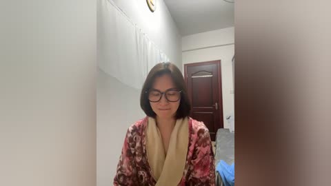 Video of a woman with shoulder-length dark hair, wearing glasses and a red floral robe, standing in a narrow, dimly lit hallway with white walls, a closed wooden door, and a blue garment on the floor.