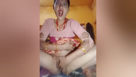 A video of a young Indian woman with fair skin, wearing a pink top and red saree, sitting on a bed, legs spread, hands holding her vulva. The background features a yellow wall and a blurry image of a man.