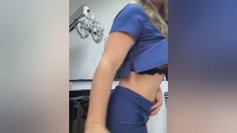 A video shows a blonde woman in a blue cropped top and high-waisted pants, standing in a medical room with an ophthalmoscope on the wall.