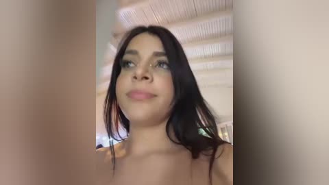 Video of a young woman with long, dark hair, light skin, and blue eyes, wearing minimal makeup, standing indoors against a blurred, beige background with wooden ceiling beams.