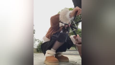 Video of a person in a white fur suit, brown fur jacket, black skirt, and tan UGG boots, crouching near a bicycle, outdoors with greenery in the background.
