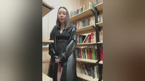 A video shows an Asian woman with long black hair, wearing a black leather trench coat, standing in a library with wooden shelves filled with books.