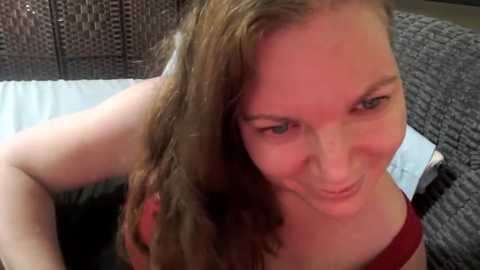 Media: A video of a young, fair-skinned woman with shoulder-length wavy brown hair, wearing a red top, smiling slightly while adjusting her hair on a dark, textured wicker couch.