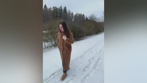 Video of a tall, slim woman with long, dark hair, wearing a brown coat and boots, standing on a snowy path surrounded by leafless trees and bushes under an overcast sky.
