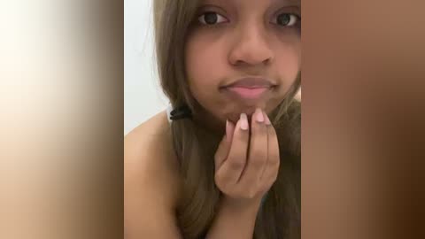 Video of a young Black woman with light brown skin, long straight brown hair, and green eyes, smiling slightly, holding her chin with her left hand.