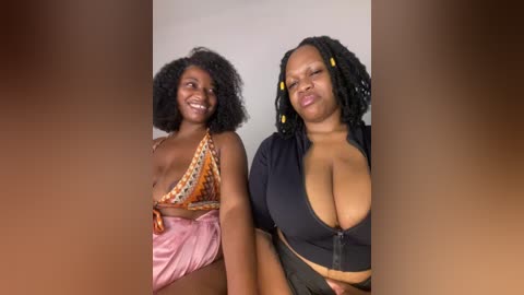 Media: Video of two African-American women with medium-dark skin tones, one in a pink satin dress with a triangle neckline, the other in a black crop top. Both have voluminous, curly hair.
