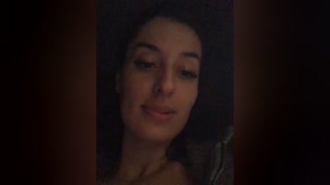 A dimly lit video of a light-skinned woman with curly hair, wearing a gray jacket, smiling slightly. The background is dark, making her face the focal point.