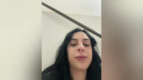 A video of a young woman with long, wavy black hair, light skin, and dark eyes, smiling slightly, standing in a minimalist room with beige walls and a metal staircase railing.