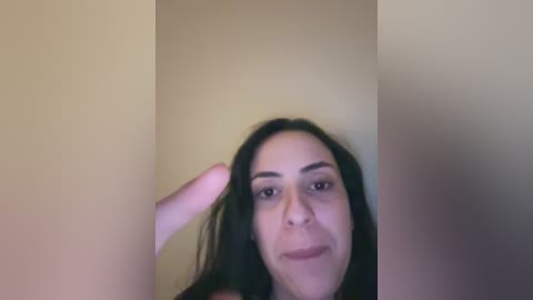 A blurry video of a woman with long black hair, light skin, and dark eyes, wearing a black top, standing in a beige room, raising her left hand.