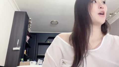 Video of a young woman with long, dark hair, wearing a white off-shoulder top, standing in a modern, minimalist bathroom with dark wooden cabinets, shelves, and a sink.