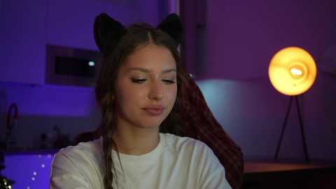 A young woman with long brown hair and cat ears, wearing a white shirt, sits in a dimly lit room with a round, glowing lamp and modern kitchen background.